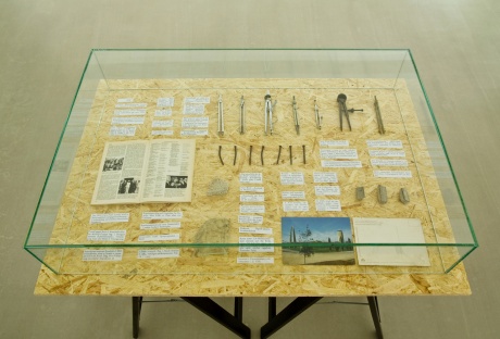 Vitrinentisch: NAILS AND DIVIDERS, 2013 |Fünf kleine Trumms/ Scherben der Niemeyer-Gebäude: drei kleine Betonteile, ein Stück Bimsbeton, ein Stück Glasscheibe und sieben Nägel (verrostet); sieben verschiedene Zirkel; Postkarte (Vorderseite und Rückseite) aus den 90ger Jahren, International Fair of Rachid Karamé, Tripoli, Foto: Alfred Moussa; Meyer’s Jahreslexikon 1975/76, Seite 82, Fotografie, Bild-Titel: Libanon. Das im Verlauf des Bürgerkriegs schwer zerstörte Geschäftsviertel Beiruts; Stein Kulturfahrplan, Ausschnitte 1975 |65 x 95 x 75 cm, Glashaube: 57 x 87 x 15 cm